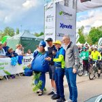 Mehrere Personen vor MSR Start-Ziel-Bogen samt Zuschauern und Kindern mit Fahrrädern im Hintergrund