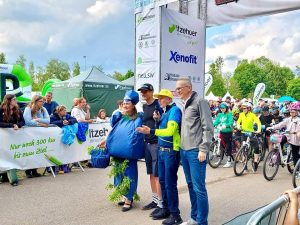 Mehrere Personen vor MSR Start-Ziel-Bogen samt Zuschauern und Kindern mit Fahrrädern im Hintergrund